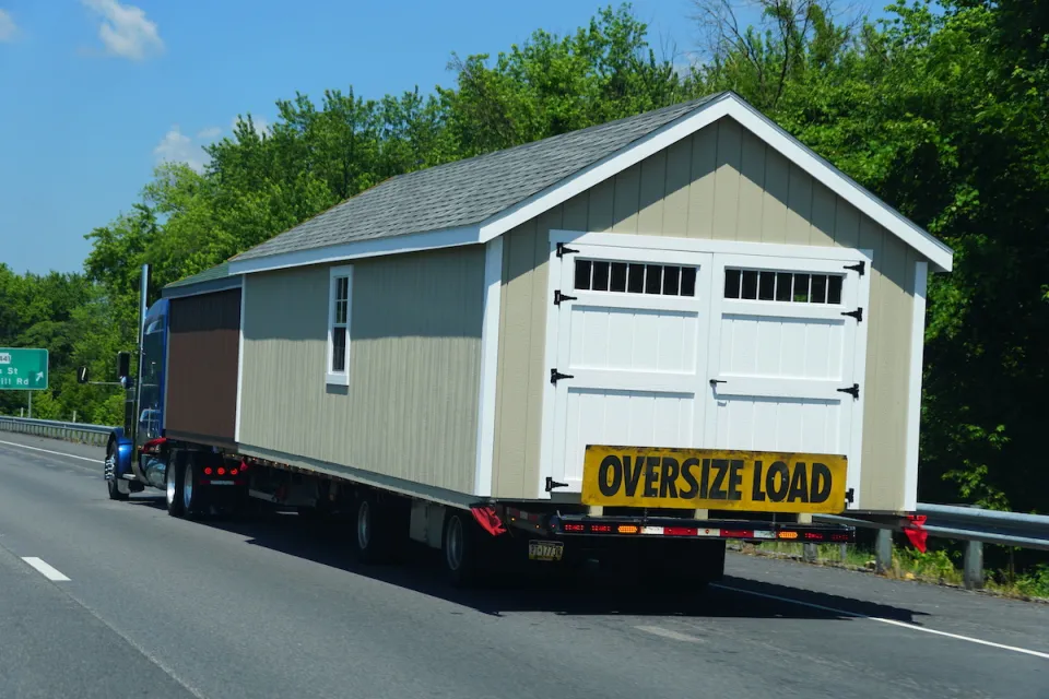 Tan modular building with white swinging barn/garage door being transported on freeway with yellow "Oversize Load" banner across back.